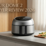 A product photograph of the unique dome-shaped Typhur Dome 2 Air Fryer, in silver and black, sitting on a modern grey concrete countertop in a well-lit kitchen next to a wooden cutting board with sliced vegetables. A stylized gold text overlay in the upper left corner reads 