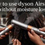 A detailed close-up photograph in a salon setting of a person wearing black gloves using a copper and silver Dyson Airstrait hair styling tool on a section of coily, dark textured hair. The tool is actively clamping the hair, which is coiling inside the tool's end, and a plume of visible steam is rising. A loose curl is forming below the tool. The background is a blurred salon counter with other products and tools. Prominent white text overlay at the top of the image reads, 